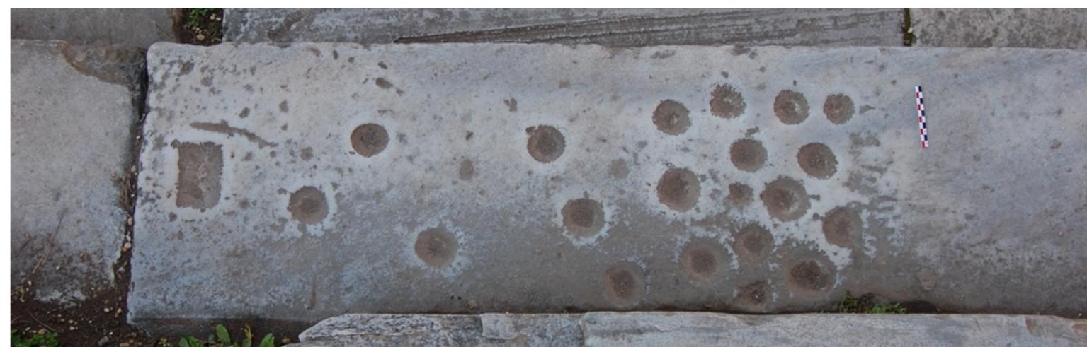 ~~ -  A clustered marble lane with twenty preserved circular pockets and one rectilinear pocket. 14 pockets of varying size and depth cluster closely together, though they are not arranged in any clear rows. Six further pockets, which likewise vary in size and depth scattered in a trail towards the opposite end of the playing surface. The final pocket, which is rectilinear, is slightly removed from the rest and is centred within the playing  surface (Schadler, 2013, 54; Schadler, 2019a, 67, figure 1; Schadler, 1994, 56). 