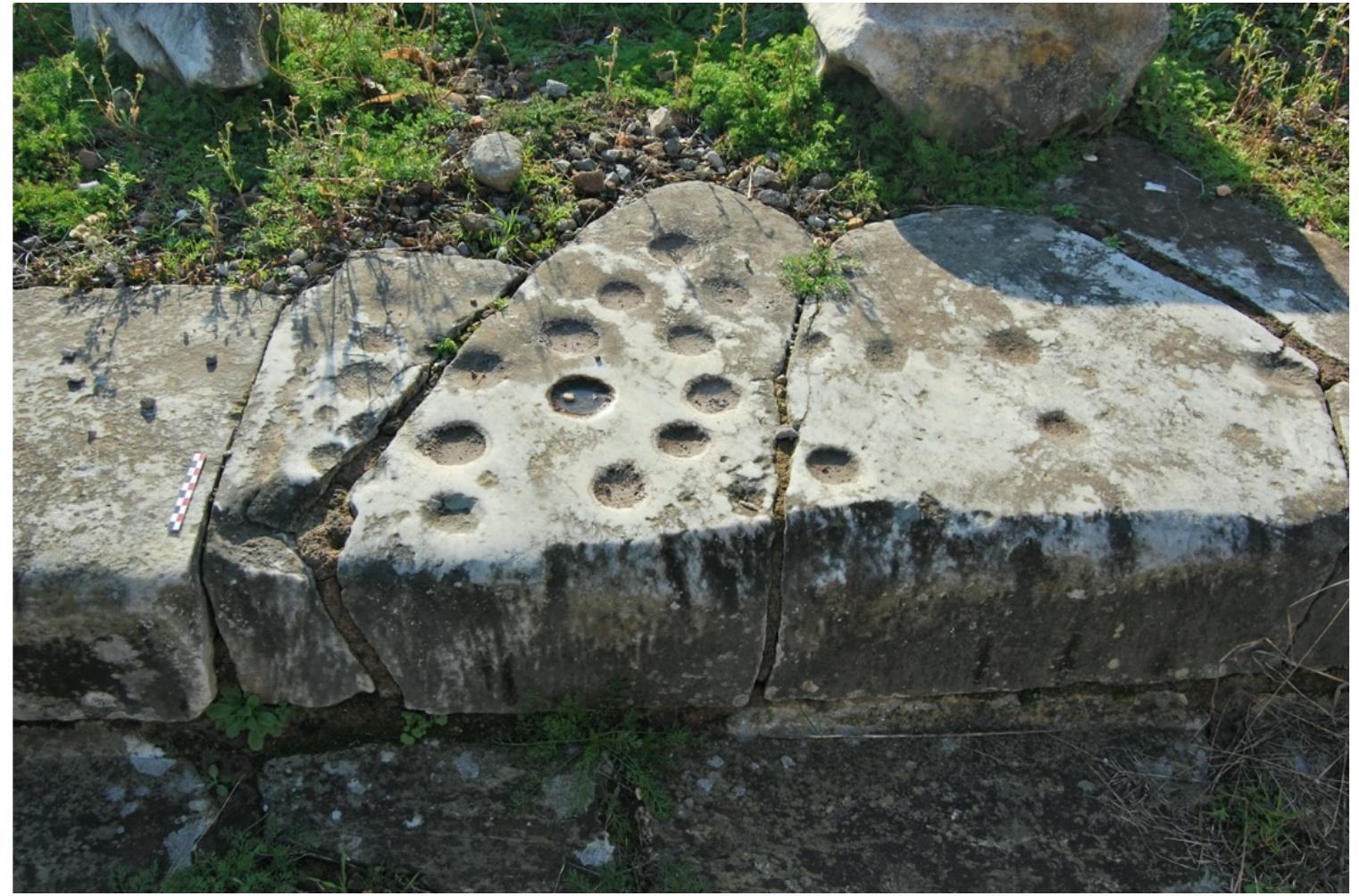 YY  A poorly preserved clustered marble lane with 18 preserved pockets(?) and a triple “starting line.” 14 of the preserved pockets cluster in a linear fashion towards the triple starting line, with two pockets bisected by the uppermost line of the triple “starting line”. Two further pockets are located well behind the triple “starting line” and the final two pockets trail away  from the main cluster and towards the opposite end of the playing surface (Schadler, 2019a, 66-67, figure 2).  in an irregular arrangement and vary in size and depth. Ihe remaining four 