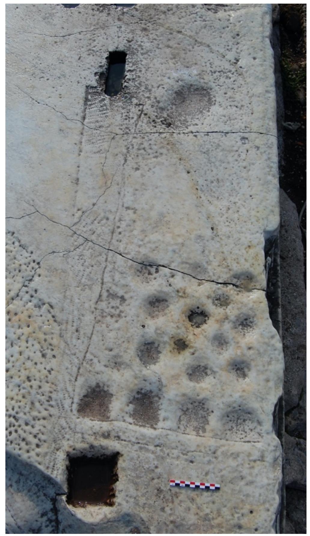 i  A partially and poorly preserved clustered marble lane with 17 preserved pockets and a double “starting line.” 16 of the preserved pockets cluster in a broadly linear arrangement in close proximity to the double “starting line.” The first four, which are closest to the “starting line” are large and shallow, while the remainder vary in size and depth. One final large, shallow pocket is located at the far end of the playing surface (Unpublished. Photograph U. Schadler). 