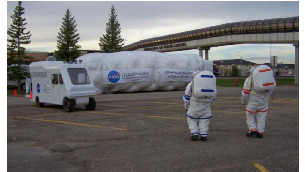 During the first week of November, 2013 three UND graduate students tested the integrated analog base elements for the first time on a 10-day mission (Figure 4). The objective of this first experience was to test the performance of the IH connected to the tunnel, the airlock interface between the NDX-2 ATs and the ER and some EVA operations with the prototypes. The crew was able to adapt to their new environment rapidly. They were able to sleep between 6-8 hours a day and take 3 meals a day. The three subjects were enthusiastic, active and well-focused. Environmental, communication and operation systems functioned efficiently. Due to weather conditions (high winds, 50 mph and freezing temperatures), there was only one minor unpredicted event during one night, that led to the partial depressurization of the IH, as the air intake was obstructed by leaves and other environmental debris.   Figure 5. UND graduate students testing the integrated analog base systems. NDX-2 ATs, Electrical Rover and Inflatable Habitat can be seen from front to back. 
