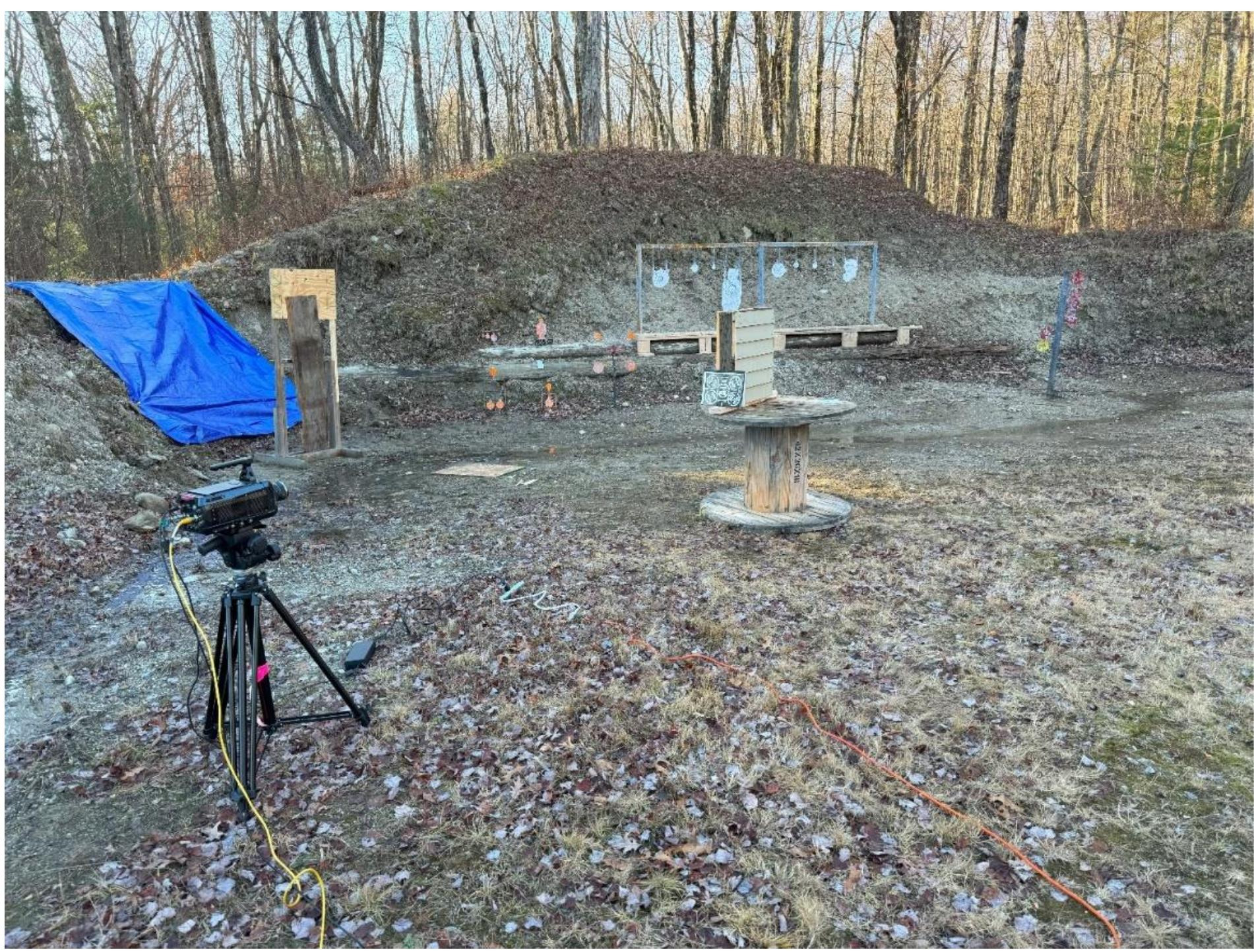; Figure 1. The high-speed camera being calibrated for a shot at a wall panel.  The high-speed camera setup consisted of a Fastec Imaging TS3 Cine camera mounted on a tripod (Figure 1) and connected by various cables to a laptop computer. The camera was remotely controlled and collected data on the shot. The camera was set to record at 2500 frames per second. The data were downloaded to the computer and field processed in Cine Viewer and saved on an external hard drive. Boxer LED spotlights with appropriate filters/gels were employed as necessary to better light the targets (Figure 2). These data were backed up on two additional external hard drives after the conclusion of the shooting day. 