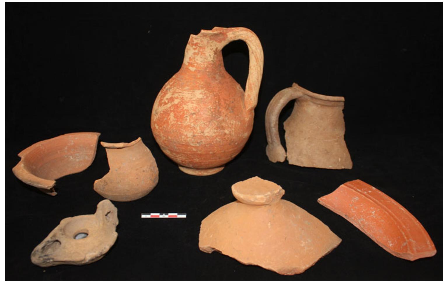 Fig. 1. Late antique assemblage from the fill of a possible cistern (photo: S. Kay).  features to the immediate east of the road. The area was chosen because of its position at an intersection of the city’s principal north-south axis of transport and movement and a secondary east-west road, as well as its potential to expose an area of potential non-elite commercial and domestic activity. Within a 15 m x 15 m trench, the secondary east—west road, paved in two different types of basalt, was found to be well worn with deep ruts and was evidently blocked in a final phase with large, reused architectural elements together with ceramic material dating to the sixth century AD. A series of small rooms lay to the south of the road, the easternmost of which had a floor of opus spicatum. Against the north-south wall extending the length of the western side of this room, a deep layer still occupied in the late sixth / early seventh century AD contained coins (ranging from the first to sixth century AD) along with quantities of bone, charcoal, gaming pieces, one die, and coloured glass. The room immediately to the west and near the centre of the trench yielded another coin (dating to the fourth century AD), reused marble architectural fragments, and contained a deep-set feature with plastered walls clearly related to hydraulic activity, perhaps a cistern, of which the fill suggests a date of mid- to late sixth century AD (Fig. 1). A small hearth built into the beaten earth floor of the room contained animal bones and charcoal, and indicated the application of intense heat. These spaces may tentatively be interpreted as a roadside taberna, or spaces generally associated with late antique commercial and domestic activities, although traces of the second to first century BC were also identified. In the western half of the trench, two distinct concrete features may reflect modest roadside features or monuments. A small extension was made on the western limit of the trench and revealed a basalt-paved section of the Via Amerina, which had suffered some modern tree-growth damage but maintained a well-preserved curb and sidewalk pavement. A longitudinal feature clearly visible in the GPR survey was revealed to be a long drain constructed from roof tiles running along the Via Amerina for the length of the trench.  