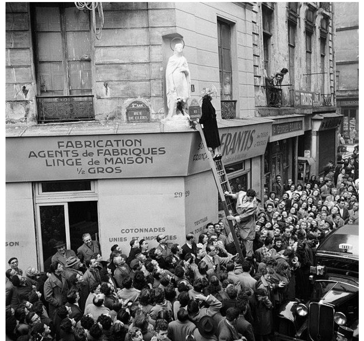 Figure 3: Decorating the Statue of Saint Catherine at the corner of the streets Cléry/Beauregard in the 9th Paris arrondissement on the 25th of November, 1950-1955, Photo taken by Willem van de Poll.  Since the Middle Ages, in Catholic France and Europe girls were under the protection  of Saint Catherine, while Saint Nicolas cared for the boys on the 6" of December. In France girls participated in devotion groups to the saint and were responsible for the confection of a beautiful headdress to "cap" her statue each year on November 25". The young women left the group when marrying, hence “capping Saint Catherine” or “coffer Ste. Catherine” became synonymous to “still being single at/after 25”. It implicated the “danger” of becoming an old undesired spinster, Saint Catherine should help finding a husband in time. 