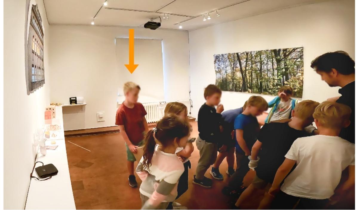 Fig. 2 - Keidel, K., Wagner, B. & Zehbe, K.-C. (2023, May 31, 00:03:30 ff., image edited). A child holds a cardboard pinhole camera while other children explore a historical collection object. Leipzig University.  Referring to the project Bildung und Objekte: Historische Sachlernprozesse in schulbezogenen Sammlungen, [Education and objects: historical general learning processes in school-related collections], such plays and games of children often arise from interacting with material objects. In the project, children have the opportunity to physically explore historical museum collection objects or their replicas. These interactions have been videographed and interpreted according to the Grounded Theory Methodology of Corbin and Strauss (2008). More spe- cifically in the terms of Eco, the collection objects in the project provide non- semiotic stimuli for children to performatively create their own meanings as well as the frameworks and rules for individual play and social games. Such plays and games and their rules are not strictly defined by the objects’ characteristics or their “affordances” (Norman, 1999) - i.e. material properties such as shapes, colours, textures or degrees of freedom of articulated objects, which would suggest a specific way of using or handling the object - but must be considered as a kind of co-creation in that the object or its properties stimulate activities. An empirical example from the project will illustrate this point.  