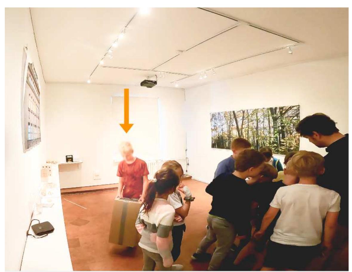 Fig. 3 - Keidel, K., Wagner, B. & Zehbe, K.-C. (2023, May 31, 00:03:30 ff, image edited). A child looks at the matte screen of a pinhole camera while other children explore a historical collection object. Leipzig University. 