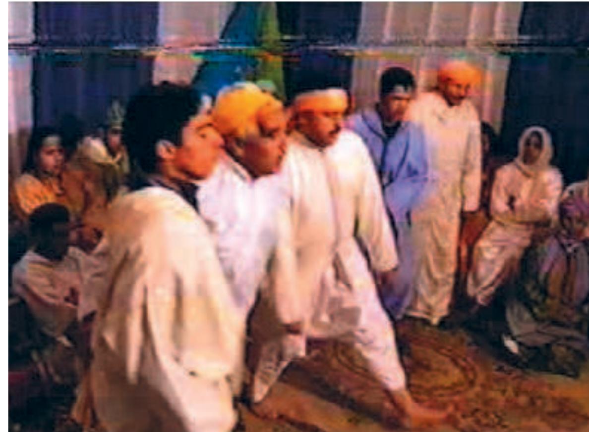 The “lions” are about to “attack” the “lionesses” in a ritual celebrated among city-dwellers (filmstill).  The singer lets his head hang and throws his body to and fro, peo: ple wail and cry out as the two hellala continue to shout lamenting verses into the room. Some of the listeners are “beaten” by the hal and along with the lion’s spirit, the heat rises in their bodies. The city dwellers watch the sacred practices unfold in their midst while the lion- dancers find their way into the trance choreography. The female lion: help them to enact and satisfy their spirits by kneeling down, trancing and also the jackal enters the scene and offers himself as sacrifice in the salon of the host.  