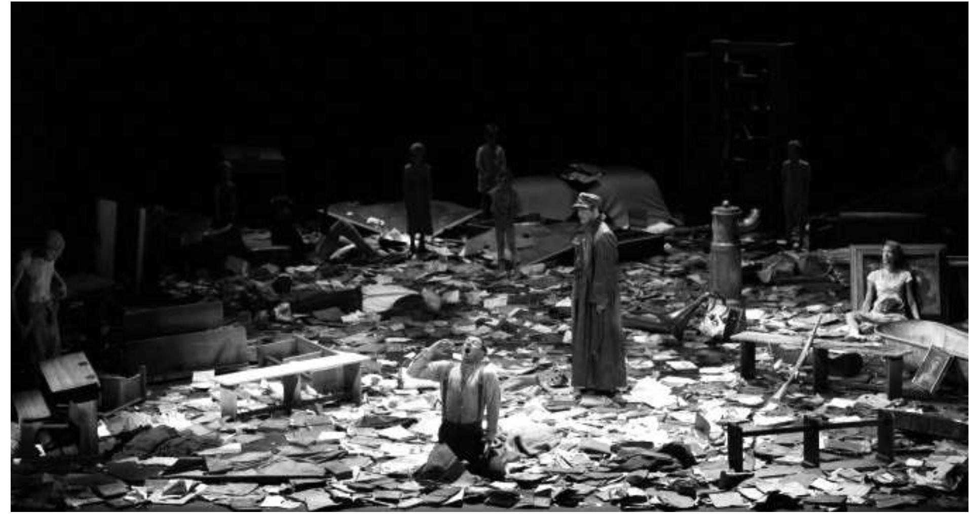 Kriesa's Galicia Act I: Norman Hacker (Lieutenant Walter), Shenja Lacher (Cadet Horvat), Ensemble in the backgroun Photo: Thomas Aurin  Act 3 begins in the same space, with white light concentrated on the big table downstage centre, where the corpse of Glembay senior is laid out covered with a white sheet; only his head and naked shoulders are visible towards the audience. Sister Angelica kneels to his right, quietly praying, while Leo sits slightly upstage to his left, drawing his father's death mask. Fabriczy, Altmann, and Silberbrandt, sit in various deep armchairs in the shadows of the periphery. After the initial stillness, the action mounts to a furious crescendo, with the corpse in the center the subject and object of more revealing recriminations, this time by the Baroness who turns Leo's defamation into her own existential truth. The staging stresses with tremendous graphic courage the horrific absurdity of this dance of death of a family, emblematic of a corrupt moribund society and epoch. The image of Sophie von Kessel straddling the corpse as she hurls her own anguish at Leo is stunning. She towers at this point above him and displays an elemental force previously unimaginable when Leo mocked her faux elegance   Galicia is based on Krieza's own wartime experience in 1916 in Galicia during the Brussilow campaign. While he did not participate in actual battles because of illness, he saw the horrendous effects of the Austrian defeat and the Russian Pyrthic victory, with one million casualties. Krieza commented, “I believe nowhere have I laughed more about human stupidity than in Galicia during 