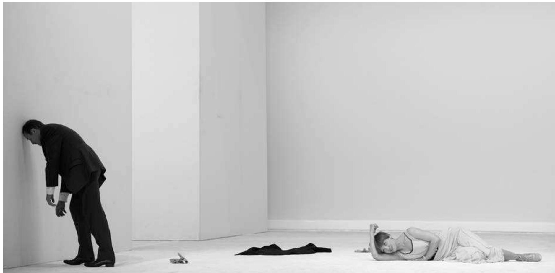 Kriesa's In Agony Act I. Photo: Thomas A urin.  The third play, without intermission like the others, is very different in texture. Set in 1922, it focuses on the intimate sphere of a dysfunctiona marriage. The totally white, totally empty set, except for a black telephone on the floor against the back wall, lit in white shadowless light came as a shock after the previous penumbral crowded sets. It laid bare quite literally the emptiness of the relationships among the three main characters and their moral and psychic disorientation. Baron von Lenbach (only mentioned in Galicia), played by Goetz Schulte, is a psychological casualty of the war and the subsequent demise of the Austrian empire—he is a drunk, a gambler, and disdains the new order where everyone has to work to eam a living. Laura, his wife, amodern assertive woman of the Glembay clan, played by Britta Hammelstein very differently from her Sister Angelica, has opened a fashion boutique and eams the money which her husband gambles away. When the play opens, Laura has just uncovered a shameful ruse by which her husband tried to trick her into giving him a considerable sum of money. He considers it her duty and the amount insignificant  e.  