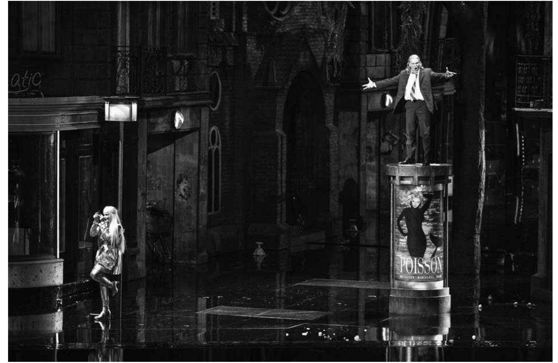 Stefan Herheim’s Rusalka at the Gran Teatre del Liceu. Photo: A. Bofill/Gran Teatre del Liceu  set in an Edwardian children's nursery. Here the events appear as part of Rusalka's imagination— her wheelchair-confined grandfather as the water gnome Vodnik, her sisters as the nymphs, her governess as the sorceress Jezibaba. Melly Still's recent and less satisfying production for Glyndebourne in 2009 refashioned Dvorak's opera as a fantasy-underwater- ittle-mermaid tale, contrasting strongly with Dvorak's stark obsession with human self-obsession.  ow Stefan Herheim's production for Barcelona's Gran Teatre del Liceu (first seen at the Theatre Royal de la Monnaie in 2008) resituates the early twentieth-century opera from an enchanted forest to a resolutely contemporary European city. For the overture willow trees sway in the evening breeze alongside a lamppost as commuters make their way home, tumbling in and out of an underground station. An endless stream of individuals run through the rainy street past a boarded up shop, church, and flickering ice-cream parlor "Lunatic": a family with a little boy, a woman carrying a violin, a blind man tapping his stick along the road, a flower seller (later shown to be Jezibaba) peddling her wares. A woman appears at a balcony. The same scene is played out as if on an endless reel, a groundhog day in which Vodnik appears trapped.   Groissbock's Vodnik the central protagonist of production. He first appears as a businessman route home; his umbrella knocked inside out the lashing wind and rain. Later, thrown out by wife— the woman on the balcony— he wanders  Herheim's — reading makes Gunther  his en by his the  streets where he is approached by Rusalka; here configured as an alluring siren-like silver-suited  prostitute with a peroxide wig. He mistreats her  but  she returns again to his abusive treatment. Camilla 