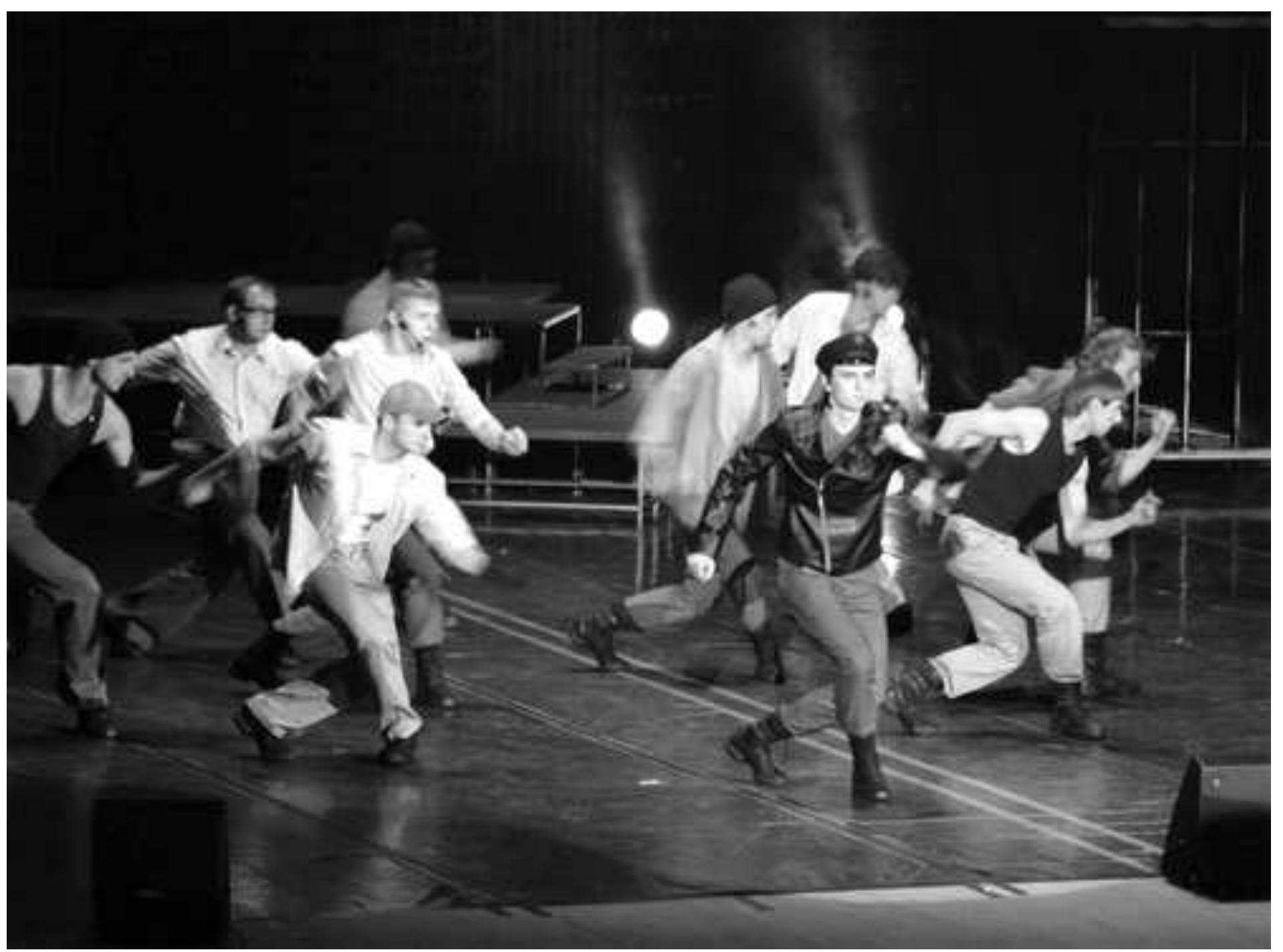 West Side Story. Photo: Courtesy of the Belarus State Musical Theatre.  The floor of the stage, now fully illuminated, is split in half by a double yellow line symbolically prohibiting passage between the two sides. The fight that ensues between two gangs, the white A merican Jets and the Puerto-Rican Sharks, is soon interrupted by the arrival of Lieutenant Schrank (Alexander Osipets). As he proceeds to badger the members of the gangs, he walks pointedly along the yellow line, which serves as the show's central metaphor. The treatment of this scene exemplifies the indirect ways in which the production suggests connections between the musical's themes and a Belarusian audience’s socio-political experience. Schrank’s mock tight-wire act is meant to convey to the young "hoodlums'" his ability to twist the law any which way he likes. Though dressed presumably as an American police officer, Osipets accentuates his character’s bullying and aggression, gesturing towards the corruption of law-enforcement agencies in post-Soviet regimes. His intonation alone makes the underlying commentary crystal clear to a Russian-speaking audience.   As the house lights dim, a black and white video of what looks and sounds like a congested avenue in the West Side of New York City in the late 1950s is projected on the backdrop, introducing the audience to the time and place of the story. In the flickering light of the screen, we can make out the outlines of the set, a jumble of metal scaffolding pushed up against the back and side walls of the stage. One by one, human silhouettes become discernible on various levels of the set. With the opening bars of "Prologue" the din of street traffic recedes; the video freezes into one still shot, then gradually dissolves.  