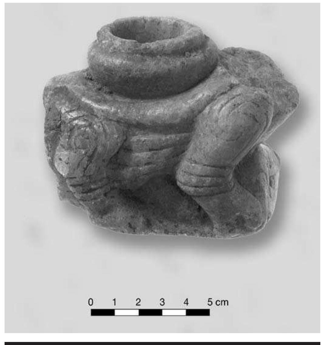 FIGURE 8.6. Feline effigy pipe (448667).  represents a canine. The tail of the feline on pipe 448667 (Figure 8.6) curves up and around the bowl, terminating near the head, in a pattern similar to that described by Brown (1976:247; 1996:513) for two puma pipes from the WPA excavations. It appears that the feline crouches over or straddles a human form. The piece is carved on all surfaces, including the bottom (Figure 8.7c). The view of the bottom clearly shows a headless human form in out- line. The person’s head appears to wrap around the front of the pipe to face out below the head of the cat. The bowl is in the form of a round, lipped pot similar to the bowl of 448663, and it sits squarely in the center of the animal’s back. The stem extends out from behind the animal, so the effigy would not face the smoker. Spirals are incised onto shoulders and knees. Lines are also incised at elbow and ankle joints. Horizontal incised lines decorate each side of the body. The animal clearly has rounded paws with incised claws and was illustrated in Hamilton (1952: pl. 17D). This pipe is manufactured from a highly crystalline stone of uncertain composition, perhaps bauxite or sphal- erite, as suggested by notes accompanying the artifact. Sediment remains in the crevice around the bowl. 