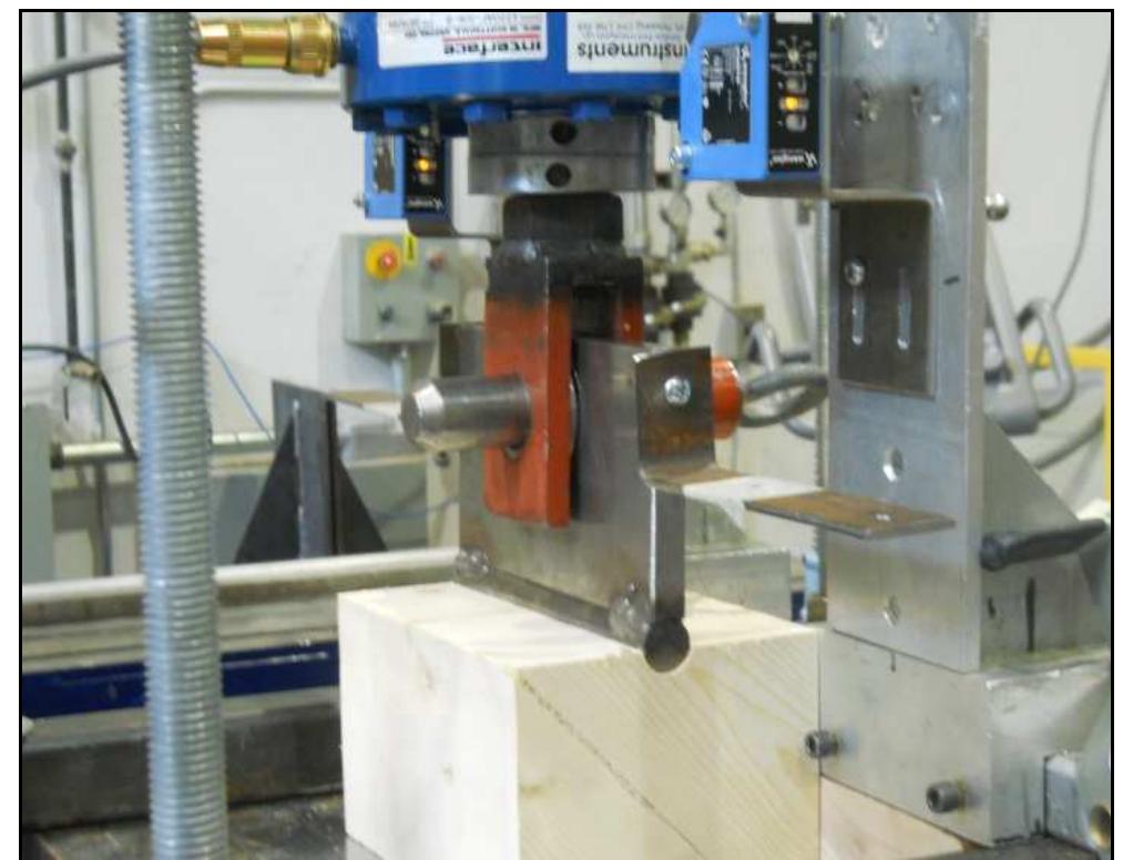 Figure 2: Embedment test set-up  The load was applied to the specimen using a fastener welded to a steel plate attached to the loading head of a hydraulic actuator (Figure 2). The specimen was loaded at a constant speed of 1.0 mm/min, and test was stopped when the load decreased to 80% of the peak load unless the displacement first reached the lesser of 7.0 mm or half the diameter of the fastener. The displacement was calculated as the average of measurements recorded using two laser sensors on the opposite ends of the specimen as shown in Figure 2. After the test, a sample was extracted from each specimen to determine the relative density and moisture content in the vicinity of the loaded zone.  