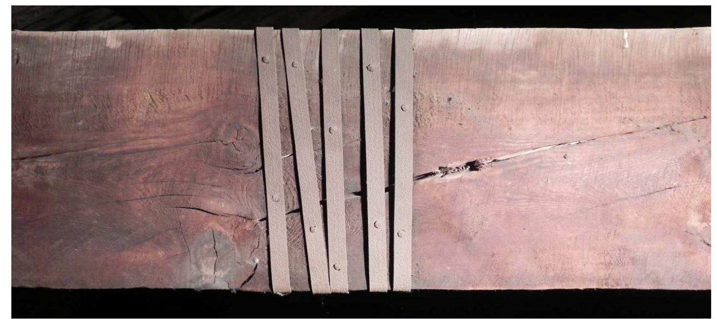 Figure 8: System used for the strengthening of the tie-beam of truss 5 (photo by E. Zamperin1).  =f.) Ue ee eee ee Ll ee lle Se ee oe Se eS Oe ee ee Sl le le  em has some peculiarity.  The tie-beams of truss 2, 4 and 5 have some big knots next to each other. This concent on of knots gives timber high local grain deviations, leading to the opening of cracks tt eaken the resistance of the tie-beam, particularly to bending moment. The three tie beat ive been repaired during time; probably truss 4 was repaired during building works, sin e materials used are similar to those used for joints between rafters and tie-beams (fig. 5 e middle and 8). Tie-beams of trusses 2 and 5 have been strengthened with a different sj m: a pair of iron bars approximately 1.5 cm thick, 5 cm wide and 170 cm long are plac slow the tie-beam in correspondence of the damage and they are kept in place with a cour ‘iron stirrups tightened with a screw and nut system. Stirrups pressing the iron bars agait nber generate a high friction that makes strengthening system effective (fig. 9). 