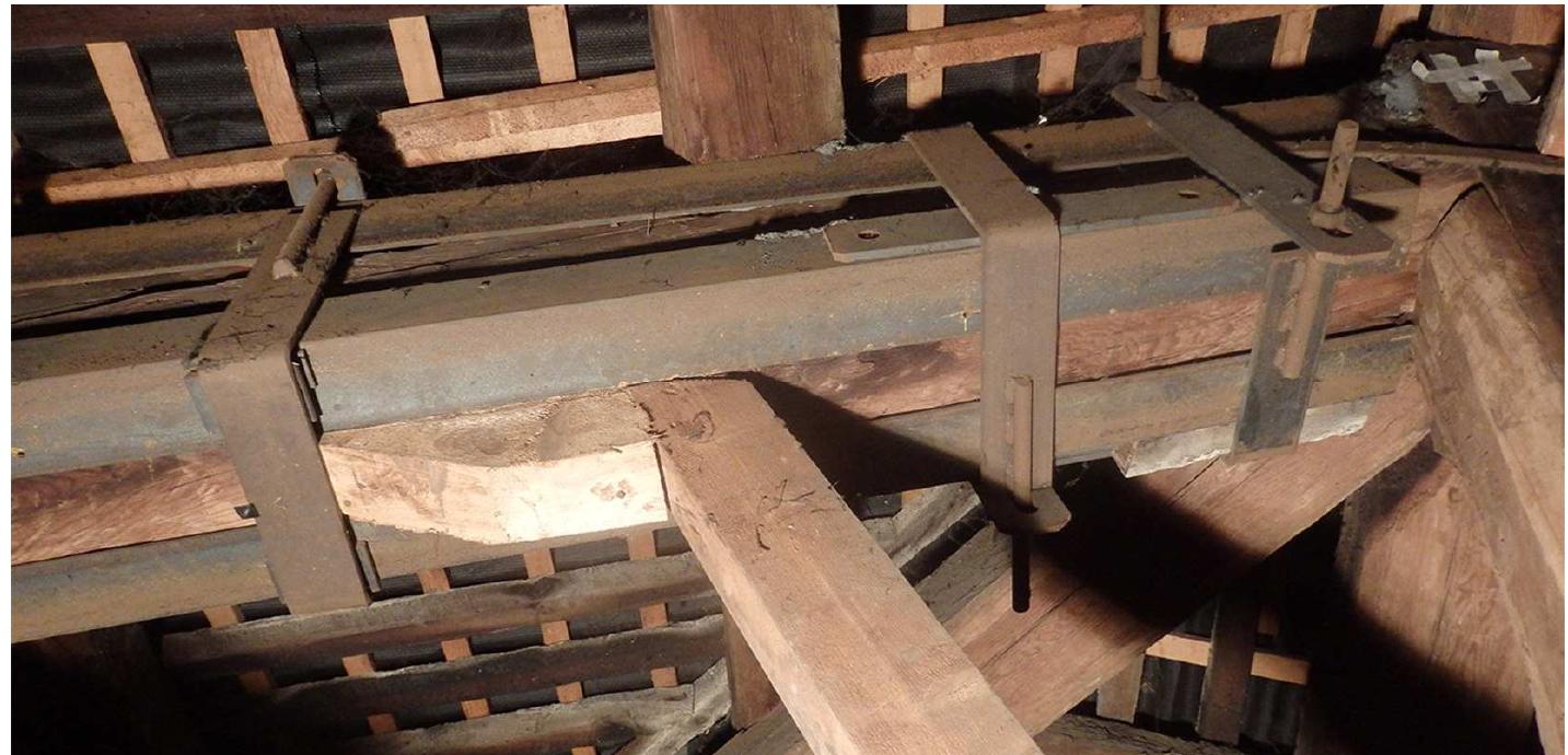 Figure 14: Strengthening system for the rafter of truss 5 (photo by M. Bosi, A. Cernuschi, E. Doria, P. Pilan)  Truss 5 has an additional peculiarity: one of the rafters has been strengthened in its upper part with four laminated steel L section bars fixed to the timber rafter with four stirrup straps. The stirrups are made of a flat laminated steel bar bent to form a U (to enclose the rafter) at whose ends short threaded rod pieces are roughly welded, and of another flat bar with a hole at each end in which threaded rod are inserted, and finally a pair of nuts to tighten (fig. 14). Simultaneously, two other struts were added in place higher than that of the original struts. 