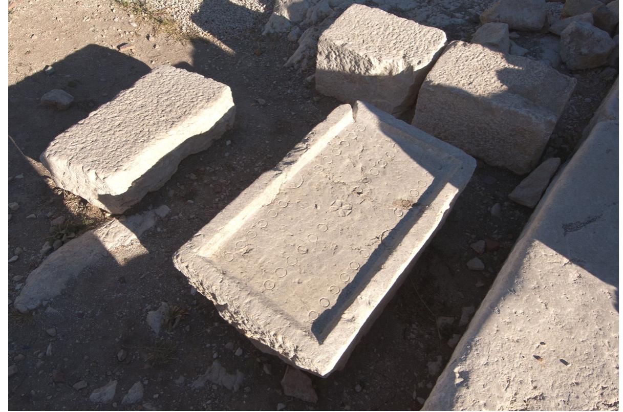 by an inscribed Maltese cross (diameter: 9cm) for the central row and two half-circles (diameter: 7cm) for the side rows.® It is flanked by the threshold of the dining hall to the west and a rectangular smooth ashlar to the east, which served as benches around the game board. Both its size and the fac that it was set up in front of a public dining hall characterise the board as a public installation, possibly even part of the furnishings of the hall itself. Moreover, this board must at least have been condoned by the ecclesiastica authorities as it was installed in a space adjacent to the staircase which lead from the square to the atrium of the city’s main church, the basilica built in the former Bouleuterion.©°  