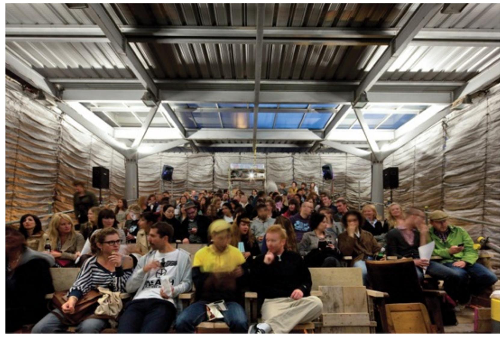 ‘actice and shifting towards a hands-on, collaborative working method, the collective yncerned with the way in which even simple, temporary constructions can act as catalysts fc ymmunity building. For example, The Cineroleum (2010) on London’s busy Clerkenwell Roz ansforms the forecourt of a derelict petrol station into a pop-up cinema. The cinema rmed by the unity of three disparate elements: the concrete and steel structure of tl ‘isting petrol station, a bank of tiered seats and an extravagant curtain of silvery draper nis billowing reflective curtain softens the concrete structure, offering both shelter and nse of theatricality to the otherwise banal site. Here the rich and ornate elements of cinerr chitecture are recreated in industrial, reclaimed or donated materials such as scaffolding ar ofing membrane, with careful attention to handmade details. The project is a celebration « e shared escapism of cinema-going: when the curtain falls, viewers are transported far fro1 e grimy site outside into a fantastic interior world.  The Cineroleum, Assemble, London, 2010. © Morley Von Sternberg 