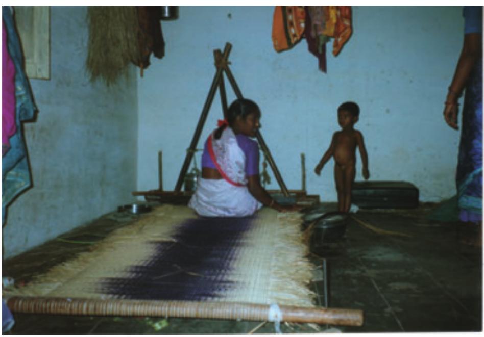 Figure 1. Regina works on a nearly completed mat.  OT  One chief difference between the loom already in use in Pattamadai and Lila’s loom is the height of the latter. Designed as a loom for a weaver to sit at on a bench (instead of on a low stool placed directly under the warp and just before the section to be woven), the new loom imposes certain changes in the layout of the mat as it is being woven (Fig. 2). Instead of the entire warp being laid out in front of the weaver so that the whole mat and its skeleton prior to weaving are visible, the warp in the modified loom is rolled up underneath the loom at the end furthest from the weaver. After the weaver weaves and packs a section of the mat, s/he rolls up the finished section underneath the warp. As the mat progresses, the rolled-up warp gradually unfolds before the weaver, is woven, compacted, and rolled up again. 