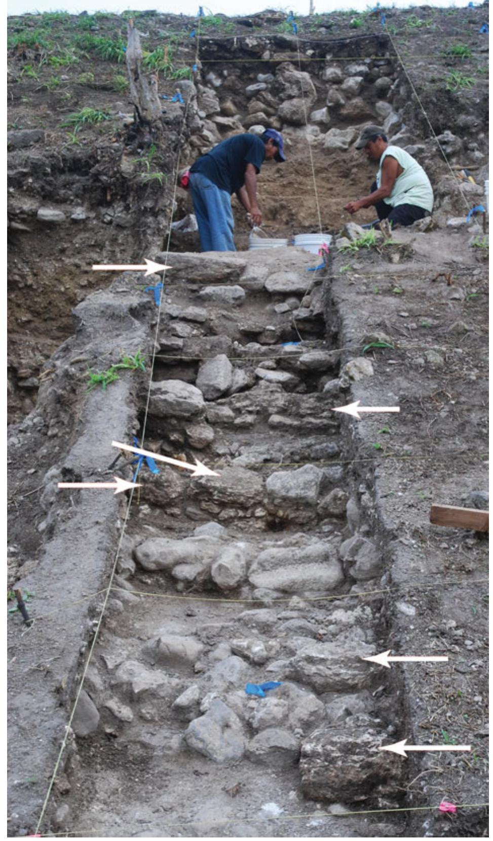 Figure 5. The terminal staircase of Structure 19a as found in excavations is seen in the foreground. The few original cut limestone blocks that formed the original stair risers were found in situ, as annotated by arrows in the photograph. All other stones are river cobble stones from the structure fill. We largely defined the staircase based on irregularly shaped river cobbles that backed the cut stone facings. Importantly, many cut stones, but not the river cobble backing, were removed. The few remaining cut stones speak to the staircase’s original engineering and the  unsystematic way in which the stones were removed. (Photograph: Lisa J. LeCount.) 