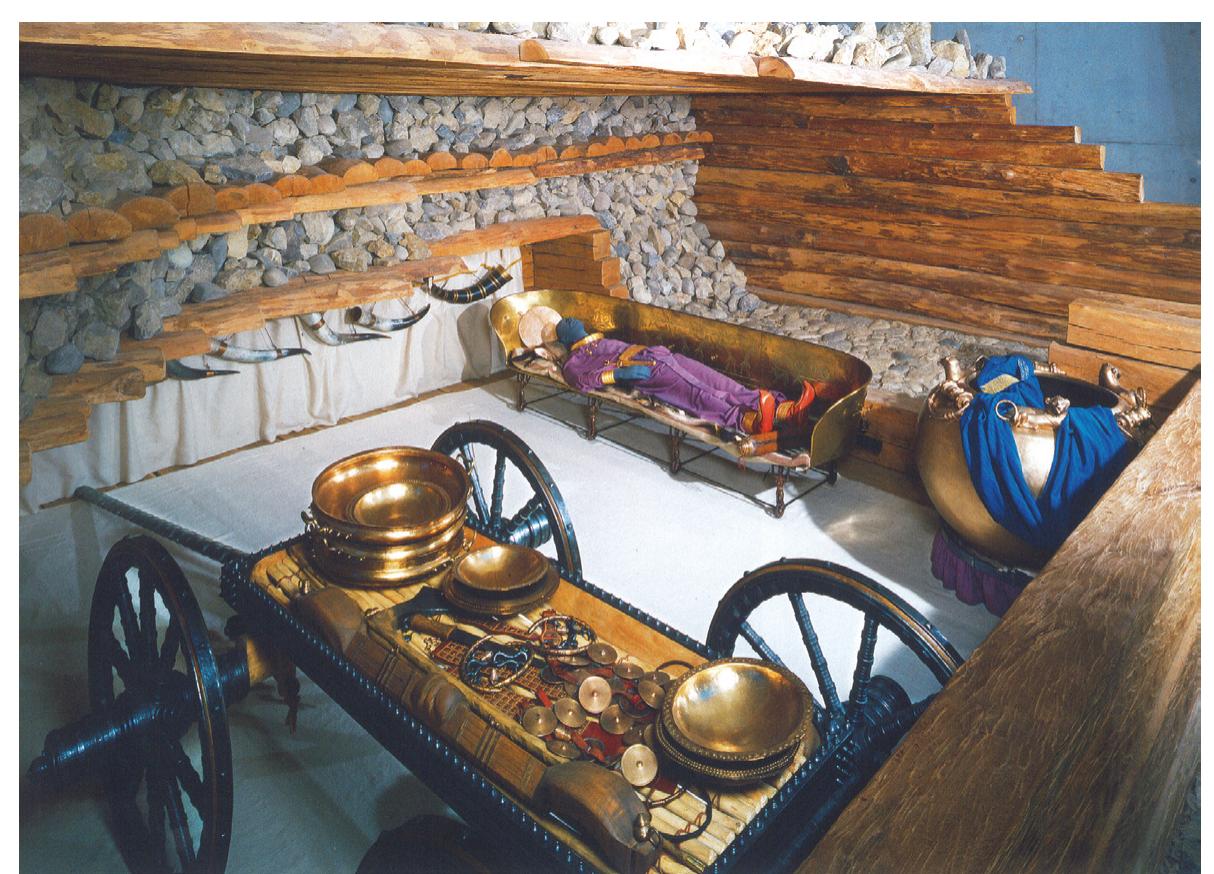 Figure 4a. Reconstruction of the ‘princely grave’ of Eberdingen-Hochdorf, around 540 BC (Hallstatt D2). The deceased man was buried in a 4.7 x 4.7m wooden chamber protected by stone packing on a kliné. Next to  the kliné was placed a bronze cauldron from Great Greece filled with  400 liters of mead. Neck and arm ring, fibulae, belts, dagger sheath and edgings of shoes were made of gold and specially for the funeral. A four- wheeled wagon, harness for two horses, as well as set of dishes such as bronze plates and drinking horns for nine persons which were used for entertainment. (Photo: S. Stork / Celtic Museum Hochdorf / Enz) 