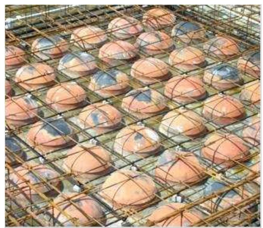 An interior view looking up at a ceiling under construction. The ceiling is a grid of concrete beams, and the square gaps between the beams are filled with reddish-brown clay tiles.