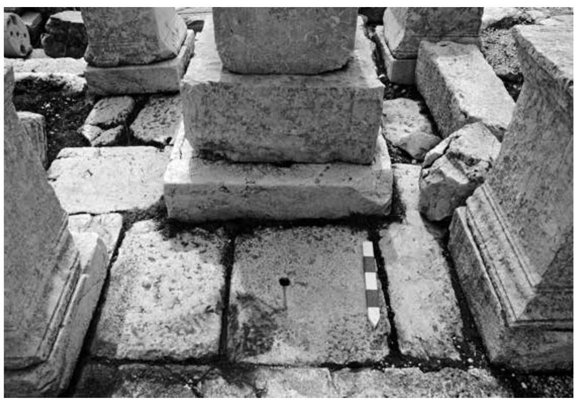 original form and position of the monument and not a late antique re-assemblage of the roof of an older structure with new pedestals as initially contended by Waelkens'’. There, a dowel hole and its pour channel have been recorded, situated in the axis of the monument, 0,40m north of the plinth of the central statue base. It indicates the place of an additional fixture, given its location most probably an altar (Fig. 6). The channel, oriented from the dowel hole towards the north, was cut into the slab over a length of 0,20m and was used for pouring molten lead to fix and seal the dowel connecting the alleged altar to the slab. This length suggests a diameter (or thickness) of ca. 0,40m for the altar. The relatively small size of the altar suggests an installation for libations or burning of incense rather than a fire altar for burning sacrificial victims”. It would then have been used for dai- y ritual practices by people frequenting the agora rather than for animal sacrifice on the occasion of civic festivals. Together with the location of the dedicatory inscription, the positioning of the altar on the north side of the monument indicates the orientation of the building towards the agora.  Crude rectangular cuttings (0,13—0,18 m wide and 0,065—0,075 m deep), are present in the top moulding of all the pedestals on the sides that face another pedestal (Fig. 7). They can be seen as indications that the sides of the structure were closed off by some kind of balustrade or fence. Barriers in shrines are generally known to have functioned as psychological rather than physical  