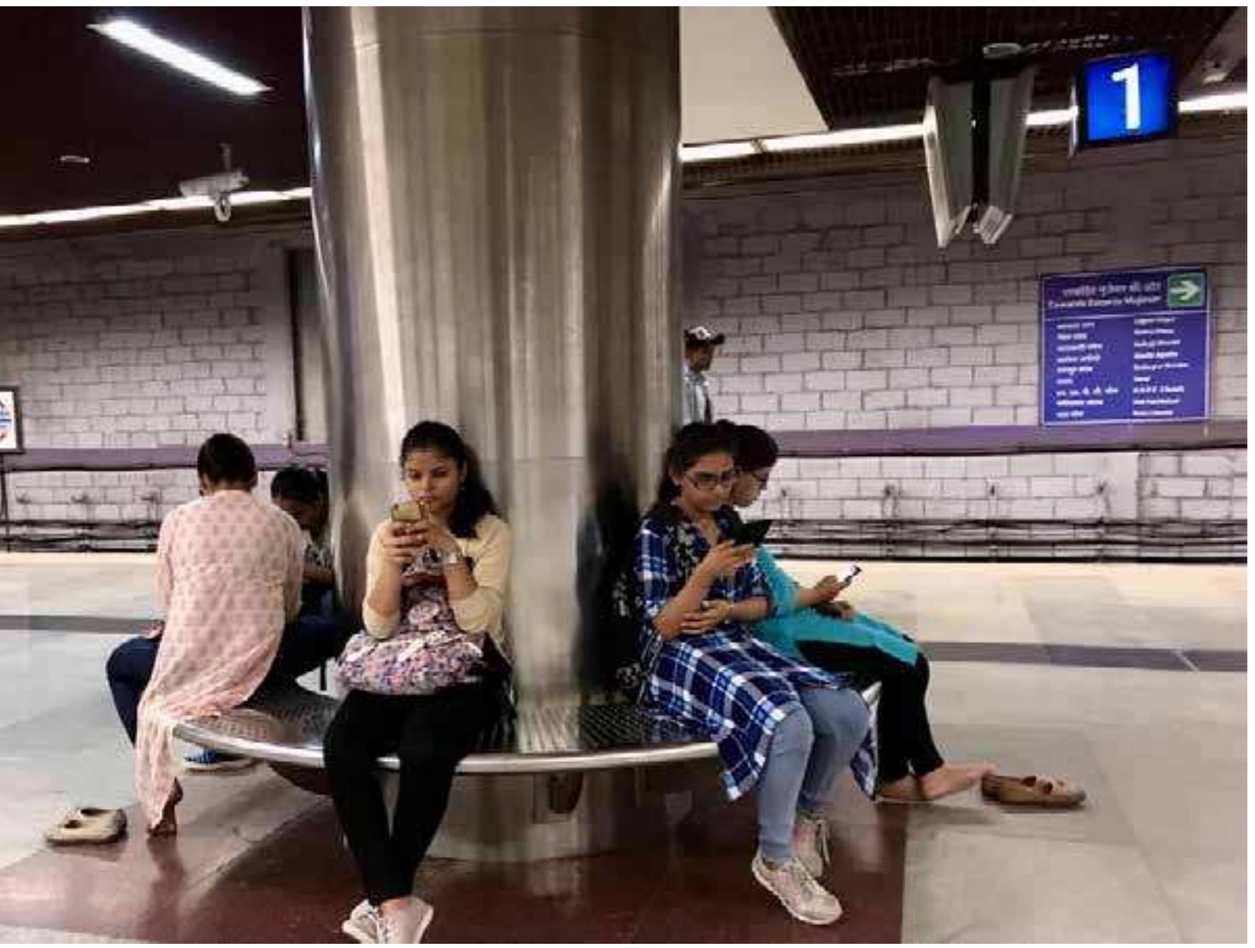 The Metro, unlike water trucks, offers no goods. It gives an energy and potentiality, a forward motion for many; but it can also emphasize a kind of stagnation. The Metro  On a Metro platform,  the comparative quiet enables phone absorption and the feeling of comfort provided by the clean floors, spurring some to slip off their shoes. Violet Line.  Photo: Rashmi Sadana, 2018.  The Metro, unlike water trucks, offers no goods. It gives an energy and potentiality, a  Outside Okhla Metro station, under the viaduct locals gather around a municip. water truck. The informal settlement here beneath the Metro lacks consistent wate infrastructure, even as state-of-the-art trains pass overhead. Most people in th neighborhood tell me they do not ride the Metro in their midst, because they do nc have the kinds of jobs — in their minds, government jobs — that would require an enable them to take it. They only step into the Metro occasionally, yet they also liv with it every day, as it cuts through their space, intones in the air. To hear versus t be heard. Many feel they have an infrastructure they do not need (the Metro), even < their water supply continues to be uneven and unpredictable.  