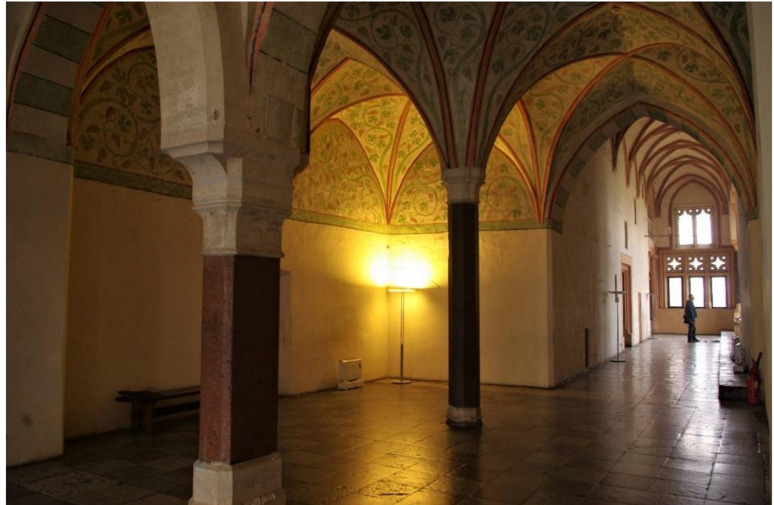 Figure 15. Malbork, Marienburg castle, Grand Master’s Palace, High Vestibule, probably 1380-1385. Photo by Tomasz Ratajczak, the author’s own work.  green, now partially hidden under a whitewash applied during restoration of the palace in the 19th century. The walls of the Summer and Winter Refectories and the High Vestibule were covered with a red-orange paint layer, while the vaults were adorned with paintings, with a dominant dark green color (Figure 15), depicting plant scrolls in two variants: the motif of acanthus leaves predominated, but some rooms took a vine motif. The interiors of the grand master’s private apartment, which were kept in a similar color scheme, were distinguished by illusionistic green curtains on a red background (Figure 16), hanging from a rod over the lower part of the walls (Herrmann 2019, pp. 293-305). They were probably more than decorative and referred to real tapestries hung on the walls of the living chambers to provide extra comfort to their users (original ornamental wrought-iron nails for hanging such tapestries have recently been identified in Siedlecin, Silesia, preserved in situ and tentatively dated to the late Middle Ages). Such a composition of painted decoration in the interior of a private room finds a direct analogy, half a century earlier, in the decoration of the papal bedroom in the Tower of Angels in the Palace of Avignon (Herrmann 2019, p. 304). The Malbork decoration, mainly abstract in character, can be dated to the last years of the fourteenth century. After a few years, it was supplemented by figural elements in the form of a gallery of grand masters in the Winter Refectory and four female saints in the grand master’s study (Herrmann 2019, pp. 295-305). Undoubtedly, he intense, almost garish colors of the most important interiors of the Grand Master’s residence left a dazzling impression of courtly splendor on the most important guests at Malbork, who were received for audiences and feasts in the palace interiors. In this way he colors breaking the monotony of brick walls became the main theme, programming  he utilitarian functions and the ideological message of the premier castle of the Teutonic Order.  