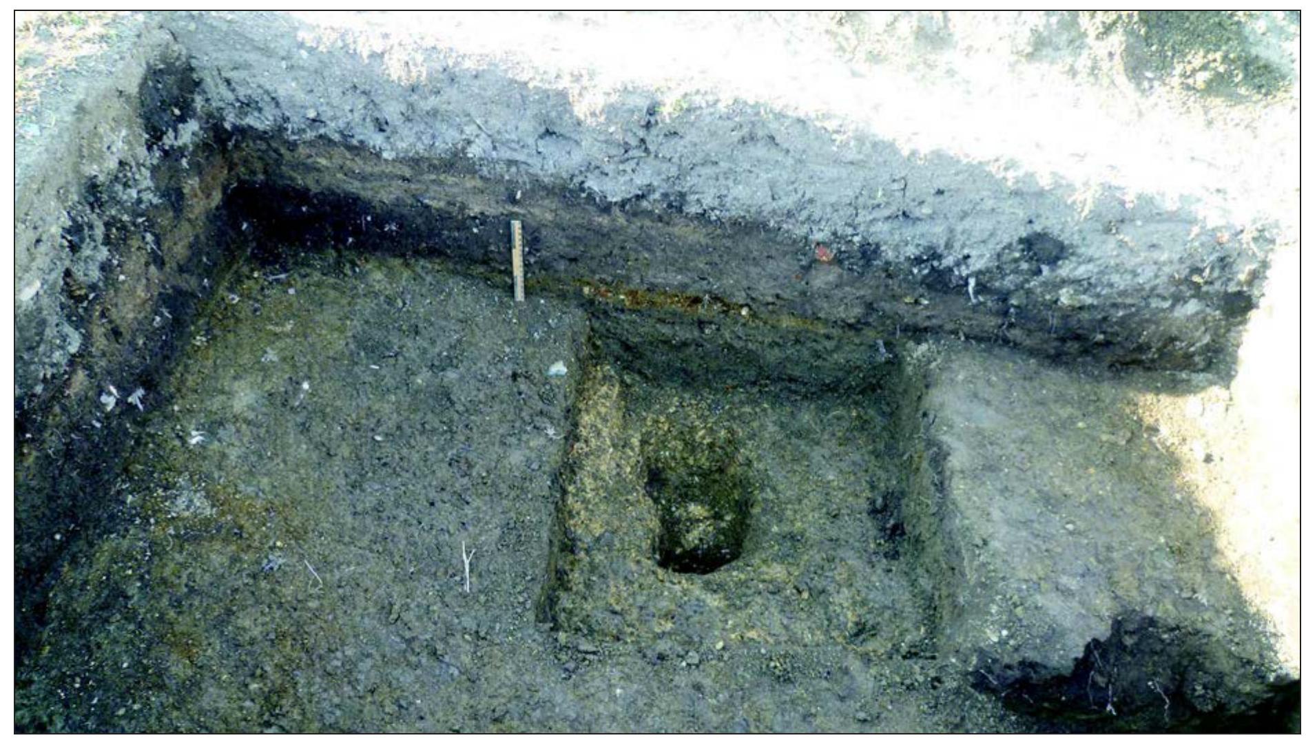 Fig. 10. Test Pit 5 (2012) on the ‘shoulder’ area showing possible gravelly surfaces on a truncated old land surface beneath midden material, with structural features cut through the base of the buried soil and into the weathered natural substrate (C. French)  A series of 11 boreholes were made across the western side of the Sana floodplain (Fig. 5). These exhibited an homogenous dark brown silty clay loam, thickening in depth from about 50cm at the base of the slope to 2m by the main road and present river channel. These soils are classified as Gleyic Fluvisols, and represent the gradual/ seasonal aggradation of overbank flood-derived deposits or alluvium. These brown silt loam to silty clay loam soils are a very valuable farming resource today and are managed by rotating from arable to grass production with a relative- ly short cycle. A few sinuous/meandering, small stream channels were evident in the farmed, for- mer floodplain area, which hint at a once more active channel system in the recent past. The fast-flowing modern river is quite deeply incised by c. 3-4m into the floodplain, and this may be its long-term course, such that the modern flood- plain represents a lengthy period of past over-  