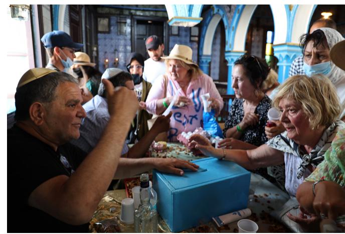Blessing of Pilgrims in the Synagogue, 2022. Photograph © Manoél Pénicaud.  Another aspect that highlights the duplicity of this pilgrimage site derives from the contrast between the austere and rigorist tendency of local Judaism, and the complex ritual choreography implemented at the sanctuary, which leaves much room for the fervour of popular devotions. According to our observation in 2014 and 2022, the synagogue becomes indeed the theatre of an intense ritual effervescence during the pilgrimage days. In the first room, a few Djerbian rabbis sitting on benches recite prayers for visitors who make offerings in exchange. These sacred gifts and counter gifts are materialized by the sharing of biscuits, dried fruits, and fig alcohol (bukha). Pilgrims mix vari- ous languages, Arabic, Hebrew, and French. 
